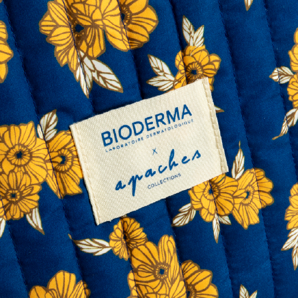 Gros plan sur l'étiquette en tissu crème avec les logos "BIODERMA" et "apaches collections", cousue sur la trousse matelassée à motif floral bleu et jaune.