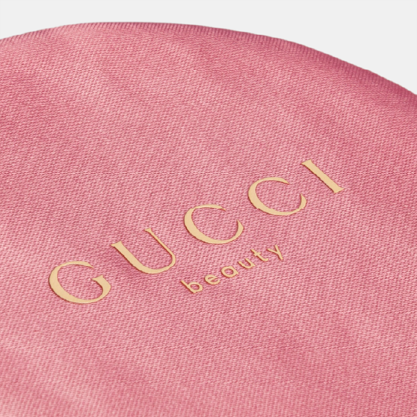Gros plan sur le logo "GUCCI beauty" en lettres dorées, délicatement appliqué sur le couvercle en satin rose au toucher velouté.