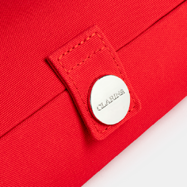 Gros plan sur le fermoir à bouton-pression argenté, gravé du logo Clarins, sur le coffret en tissu rPET rouge.