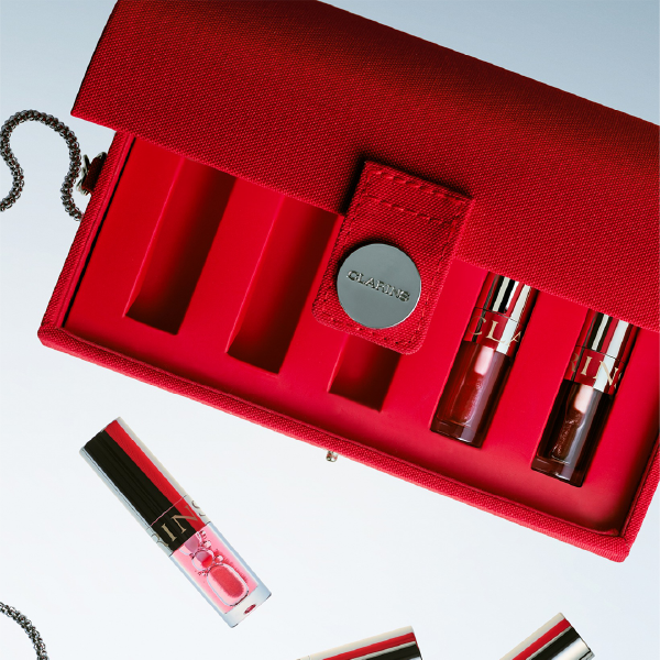 Vue de dessus du coffret rouge Clarins ouvert, montrant les compartiments de rangement avec deux huiles à lèvres à l'intérieur et d'autres produits posés à côté.