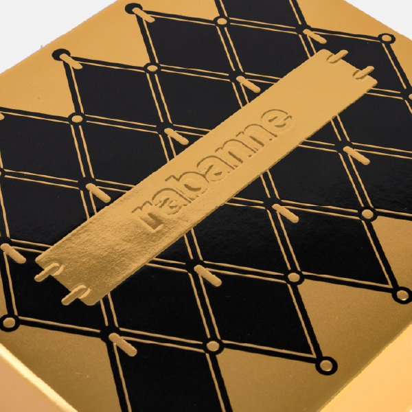 Gros plan en angle sur le couvercle du coffret, mettant en évidence le logo "rabanne" embossé sur la plaque dorée et le motif graphique noir.