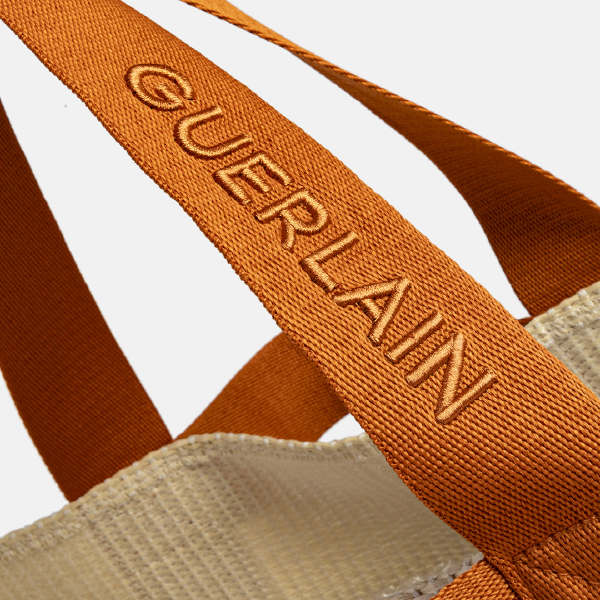 Gros plan sur l'anse en rPET effet coton terracotta du sac Guerlain, montrant le logo "GUERLAIN" en broderie en relief ton sur ton.