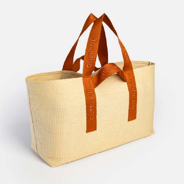Vue de trois-quarts du sac de plage Guerlain, montrant sa forme cabas et sa structure en Paperstraw léger et durable.
