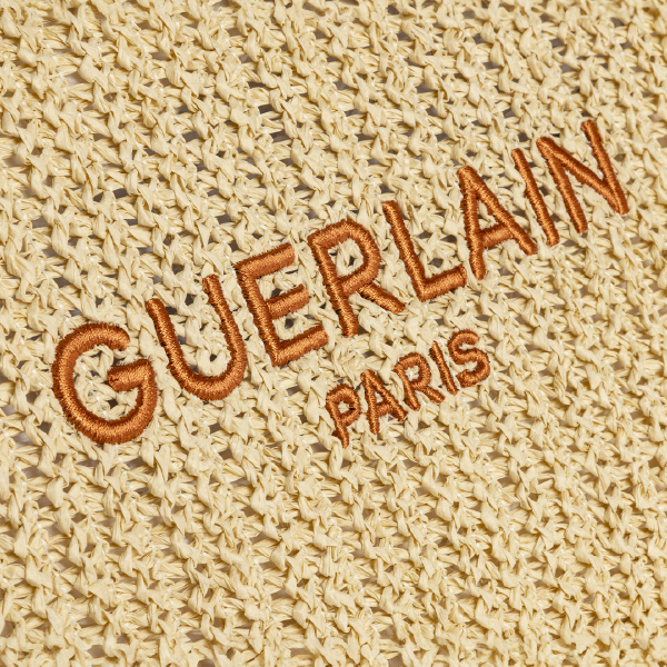 Gros plan sur la broderie "GUERLAIN PARIS" sur la trousse, mettant en valeur la texture du Paperstraw tressé.