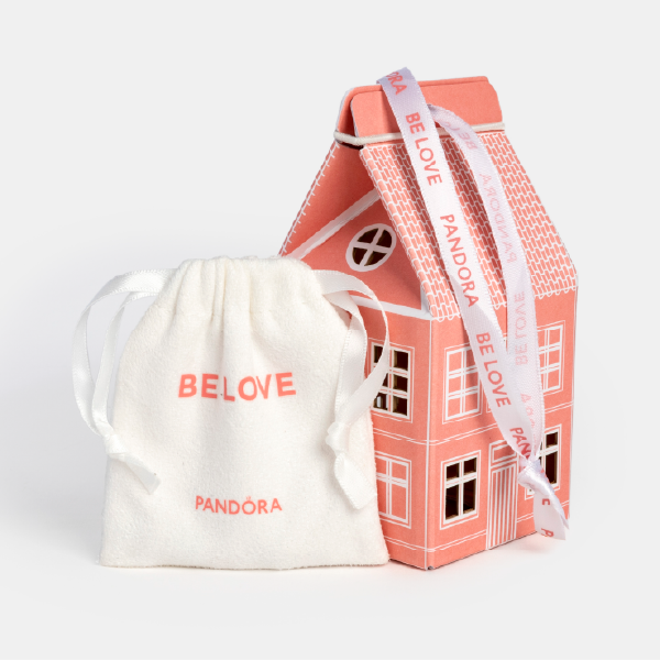 La maisonnette en papier rose fermée, posée à côté de son pochon en tissu blanc siglé "BE LOVE" et "PANDÓRA".