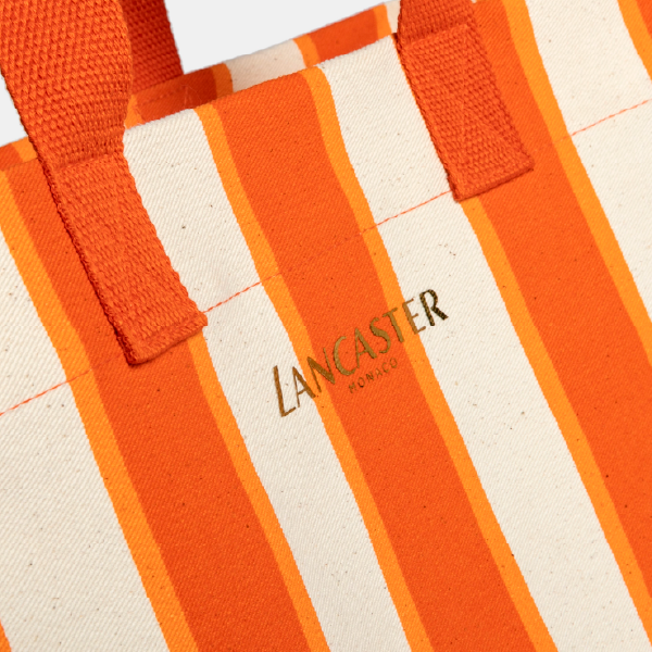 Gros plan sur le logo doré "Lancaster Monaco" sur le sac de plage en twill de coton biologique certifié GOTS, avec ses rayures orange.