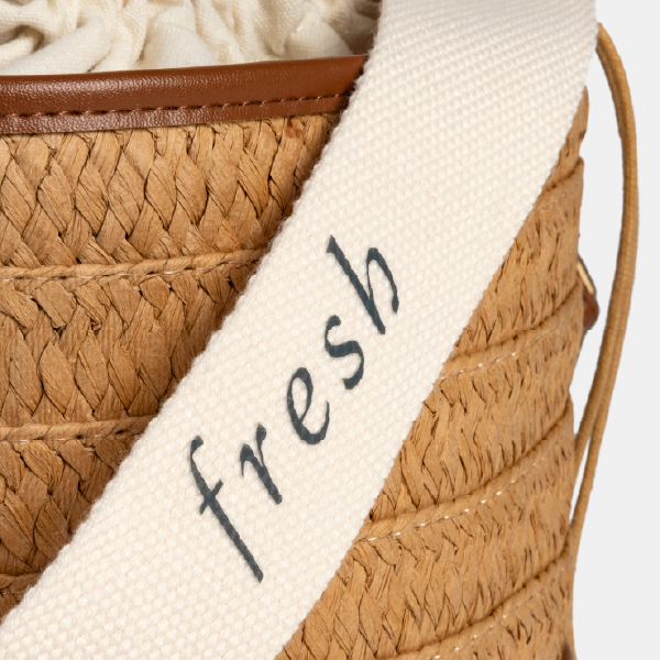 Gros plan sur la anse en coton blanc du sac seau, avec le mot "fresh" inscrit, montrant la texture du tissage sur le fond en paille de papier.