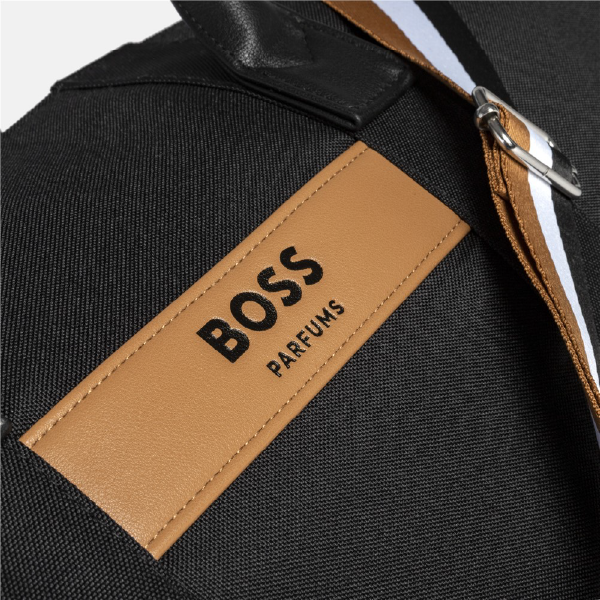 Gros plan sur le patch en PU camel du sac Hugo Boss, avec le logo "BOSS PARFUMS" embossé, et la sangle tricolore signature visible à côté.