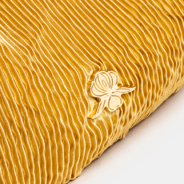 Gros plan sur le monogramme en métal doré en forme d'orchidée, signature de Sisley, posé sur le satin plissé jaune du sac.