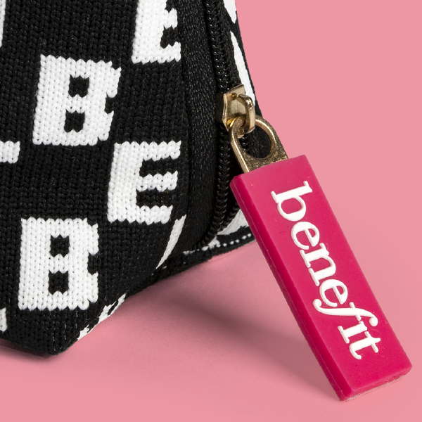 Gros plan sur la tirette de zip rectangulaire rose vif siglée "benefit", attachée à la fermeture éclair de la trousse en maille.