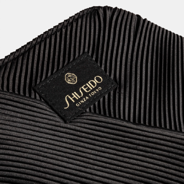Gros plan sur le patch en tissu noir avec le logo Shiseido Ginza Tokyo doré, cousu sur la texture plissée de la trousse.