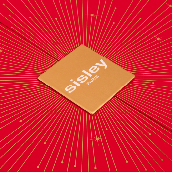 Gros plan sur la plaque carrée dorée au centre du coffret cœur Sisley. Le logo "sisley PARIS" est imprimé en blanc, sur un fond de lignes dorées rayonnantes.