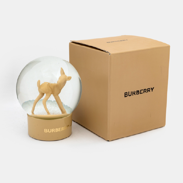 La boule à neige Burberry avec sa figurine de faon posée à côté de sa boîte de présentation cubique de couleur beige, sur laquelle est inscrit le logo Burberry.