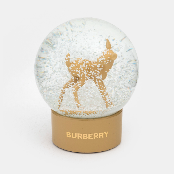 Boule à neige Burberry avec des paillettes blanches imitant la neige qui tombe autour d'une figurine de faon. Le socle beige est marqué du logo Burberry en doré.
