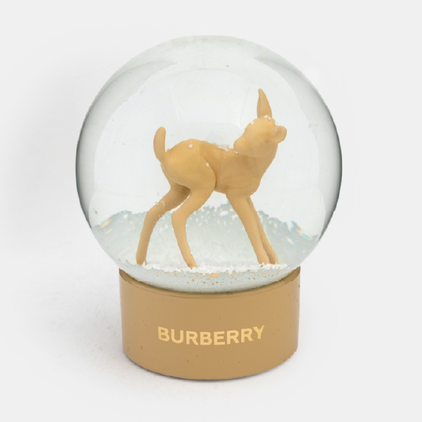 Vue claire de la boule à neige Burberry montrant la figurine de faon en résine sur un lit de neige, avant que la neige ne soit secouée. Le logo doré est visible sur le socle.