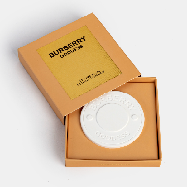 Vue de dessus de la céramique à parfumer Burberry Goddess dans son boîtier ouvert. Le médaillon blanc en relief repose dans la boîte couleur sable, avec son couvercle posé dessus.