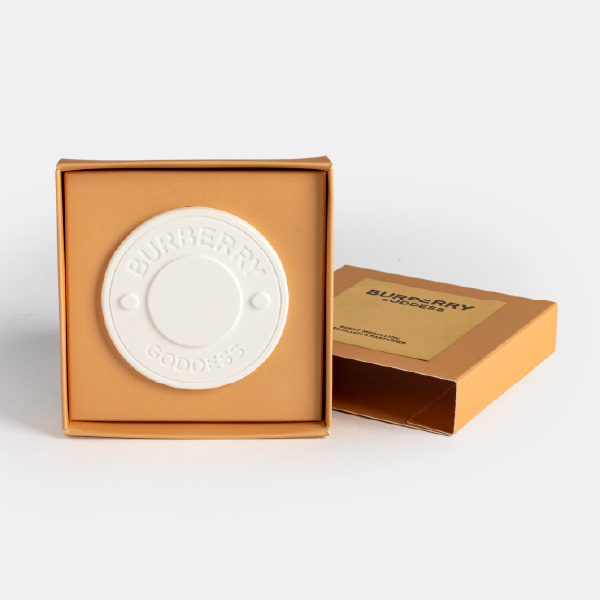 Le médaillon en céramique blanche Burberry Goddess présenté dans le fond de sa boîte, avec le couvercle de luxe posé à côté sur fond blanc.
