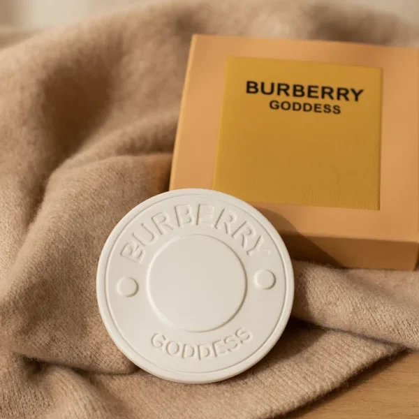Céramique à parfumer Burberry Goddess, ronde et élégante en blanc embossé, présentée sur un tissu doux avec son coffret beige et jaune en arrière-plan.