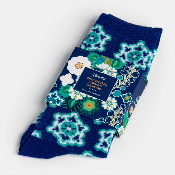 Paire de chaussettes bleues à motifs ski rétro, pliées et maintenues par un étui en carton au design floral et coloré signé Catalina Estrada.
