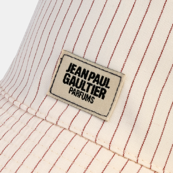 Gros plan sur l'étiquette tissée du bob, avec le logo "JEAN PAUL GAULTIER PARFUMS" en noir sur fond crème, cousue sur le tissu à rayures.