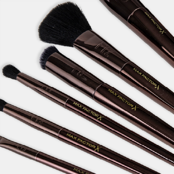 Gros plan sur plusieurs pinceaux Max Factor X, mettant en évidence le logo doré sur les manches couleur bronze et les viroles métalliques.