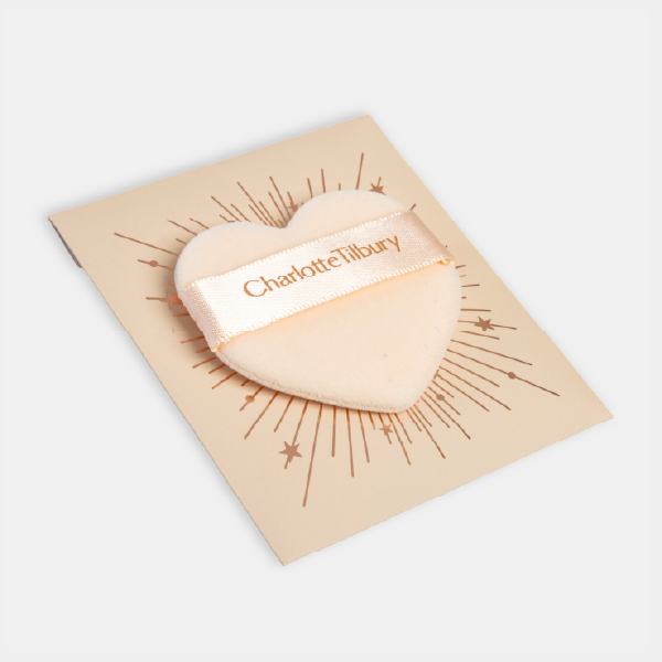 La houppette en forme de cœur Charlotte Tilbury posée sur sa carte de présentation, décorée d'un motif de cœur rayonnant.