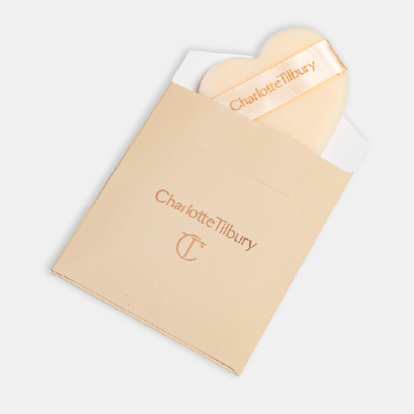 La houppette en forme de cœur Charlotte Tilbury qui sort de sa pochette en papier assortie, révélant son ruban siglé.