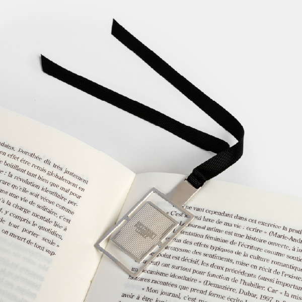 Mise en situation d'un marque-page en métal Gucci utilisé dans un livre ouvert, montrant son design de flacon et son ruban en gros-grain noir.
