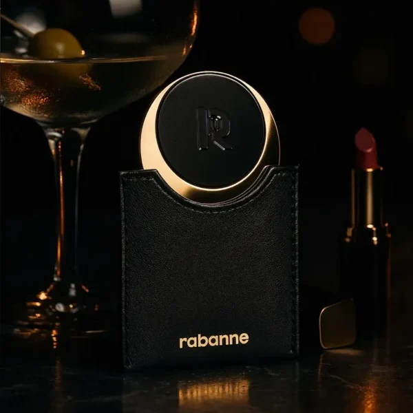 Miroir de poche Rabanne au design élégant noir et or, présenté dans un étui en cuir avec logo doré, accompagné d’un verre de cocktail et d’un rouge à lèvres en arrière-plan pour une mise en scène luxueuse et raffinée.