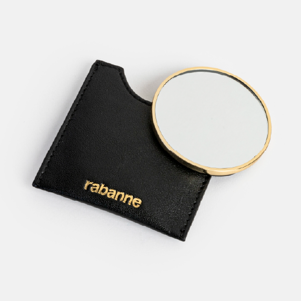 Le miroir de poche rond Rabanne posé à côté de son étui noir, montrant la surface réfléchissante et le contour en métal doré.