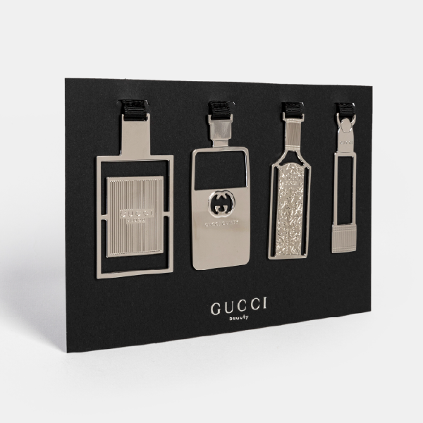 Ensemble des quatre marque-pages en métal Gucci Beauty, en forme de flacons de parfum et de rouge à lèvres, présentés sur leur support noir.